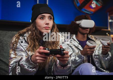 Giovane donna e amico anonimo in occhiali VR seduti sul divano insieme e giocare a videogame con joystick a casa Foto Stock