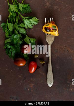 Una forchetta con una pasta di conchiglione ripiena di pomodori ciliegini, olive nere e capperi su un asse di metallo arrugginito. A lato: Prezzemolo fresco. Foto Stock