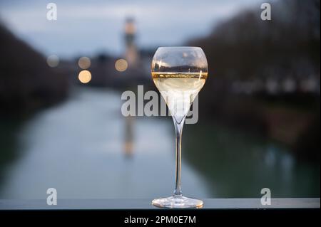 Bicchiere di vino bianco frizzante francese con champagne bolle all'aperto con vista sulle acque verdi del fiume Marne e le luci del viale Promenade in mor Foto Stock