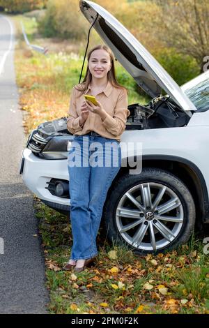 Una donna attende assistenza vicino alla sua auto rotta sul lato della strada. Donna con un'auto rotta con cofano aperto. Turbato giovane donna con cellulare vicino Foto Stock