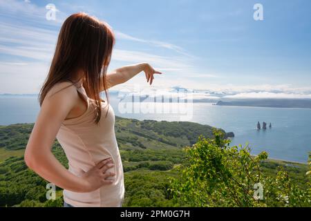 Donna senza volto punta con la mano al mare blu sotto, in piedi in alto in montagna contro la vista panoramica della foresta verde e dell'oceano. Donna irriconoscibile Foto Stock