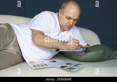Un uomo d'affari si trova su un divano e guarda le tendenze dei dati su grafici e grafici in un tablet, prende appunti in un notebook. Lavoro a distanza Foto Stock