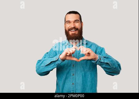 L'uomo allegro bearded sta facendo il gesto del cuore sopra lo sfondo grigio chiaro in uno studio. Foto Stock