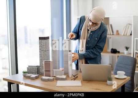 Donna d'affari musulmana professionista in hijab tradizionale che prende le misure dei modelli della costruzione usando il righello durante la giornata lavorativa in ufficio. Designer creativo che controlla la precisione dei dettagli tramite laptop. Foto Stock