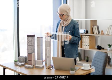 Donna musulmana gioiosa in hijab e occhiali che tengono un tablet moderno mentre prende modello in scala di grattacielo multi-piano dalla scrivania. Buon designer che controlla il disegno digitale in un ambiente di lavoro creativo. Foto Stock