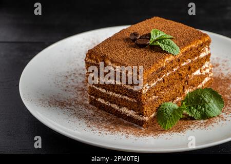 Tiramisù dessert italiano su sfondo scuro. Fetta di torta di caffè su piatto bianco. Dolce vegetariano decorato con foglie di menta. Foto Stock