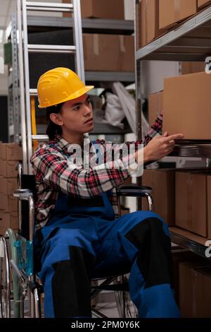Responsabile delle spedizioni asiatico con ordine di prelievo per invalidità in magazzino industriale. Addetto al magazzino dei servizi postali che mette la scatola di cartone sullo scaffale, lavorando in un ambiente di lavoro inclusivo Foto Stock
