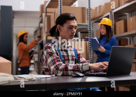 Addetto al servizio postale che crea la fattura del pacco sul laptop mentre lavora in magazzino. Storehouse asiatico uomo dipendente a digitare sul computer mentre fare la gestione di inventario delle merci Foto Stock
