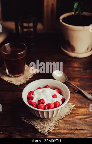 Ciotola di Dessert estivo con lamponi maturi e yogurt greco e bicchiere di caffè su superficie di legno Foto Stock