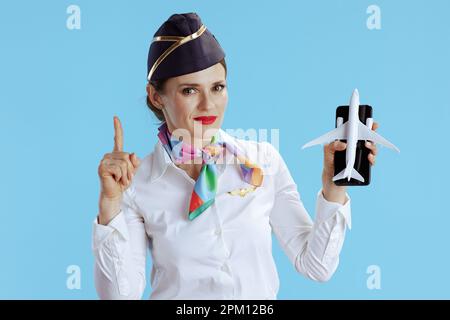 donna sorridente moderna di gestione su sfondo blu in uniforme con un po 'di aereo e smartphone con dito sollevato attirare l'attenzione. Foto Stock