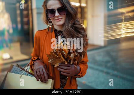 Ciao ottobre. giovane donna in trench marrone con borse da shopping e foglie gialle autunnali vicino al negozio in città. Foto Stock