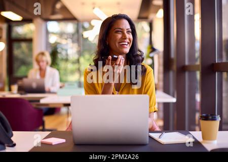 Donna d'affari matura che lavora su laptop alla scrivania in ufficio parlando con il microfono del telefono cellulare Foto Stock
