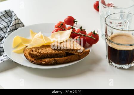 Piatto della colazione con formaggio gouda, pane, pomodori e un bicchiere di caffè fresco su tavolo bianco, primo piano Foto Stock