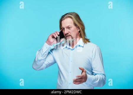 Uomo d'affari fidato e maturo che ha una conversazione seria sul telefono cellulare in piedi su sfondo blu Foto Stock