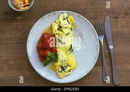 Omelette fatta in casa con spinaci e pomodori ciliegini arrosto in piatto di ceramica su tavolo rustico in legno, sana colazione omelette francese con verdi, direcam Foto Stock
