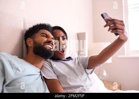 Giovane coppia a casa in posa per Selfie sui telefoni cellulari in Bed Together Foto Stock