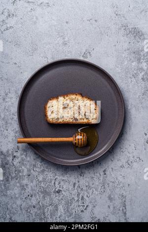 Fetta di pane dolce con noci e miele cucchiaia su piatto di ceramica su sfondo grigio vista dall'alto Foto Stock