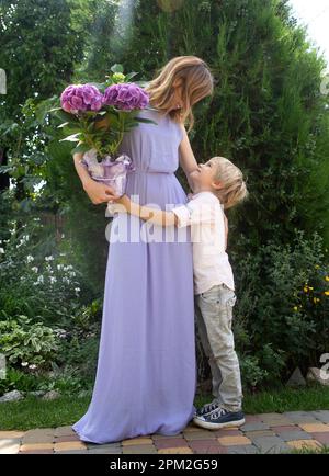 Famiglia felice, maternità gioiosa. Una donna vestita con un'ortensia in una pentola e un ragazzo che abbraccia il suo stand in giardino. Congratulazioni da figlio per m Foto Stock