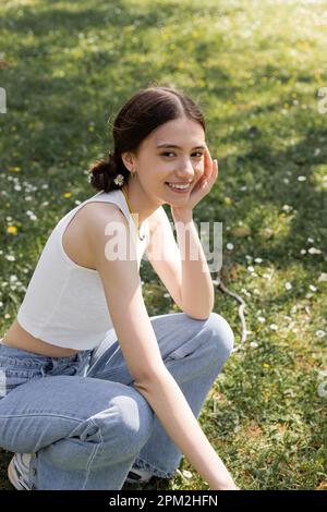 Giovane donna sorridente con fiore nei capelli guardando la macchina fotografica sul prato in parco, immagine stock Foto Stock