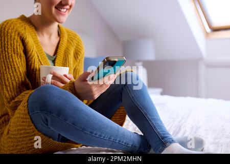 Primo piano della giovane donna seduta sul letto a casa sorridente guardando il telefono cellulare tenere bevanda calda Foto Stock