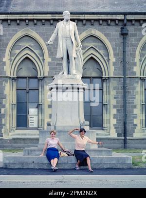 Un'immagine storica del 1981 di due giovani donne che sventolano e suonano alla base della statua di William Rolleston all'esterno del museo di Canterbury, nella città di Christchurch, sull'isola meridionale della Nuova Zelanda. Nel terremoto di Christchurch del 2011, la statua è stata rovesciata dal suo zoccolo ed è stata danneggiata, ma è stata riparata e reintegrata nel 2016. Foto Stock