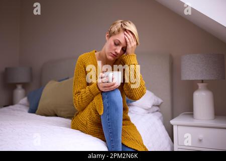 Giovane donna infelice con bevanda calda seduta sul letto a casa Foto Stock