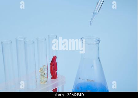 Concentrarsi sulle gocce con soluzione blu che gocciola da una pipetta graduata in un matraccio da laboratorio e sulle provette con capsule di gel Foto Stock