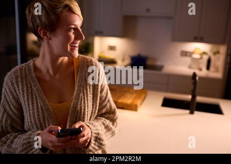 Sorridente giovane donna in salotto a casa guardando il telefono cellulare di notte Foto Stock