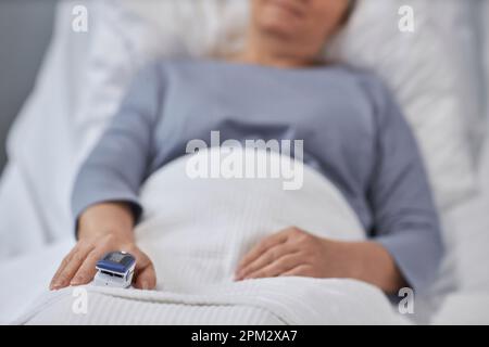 Primo piano della paziente anziana sdraiata sul letto e che esamina la sua pressione sanguigna con un dispositivo speciale sul dito Foto Stock