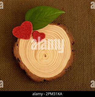 Sfondo marrone scuro iuta dove c'è un pezzo di legno tagliato dove si possono vedere i suoi cerchi, due cuori rossi con glitter appaiono e una foglia verde viene Foto Stock