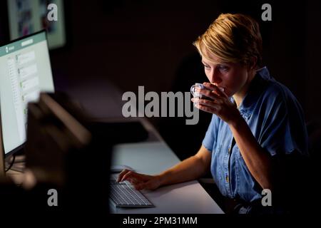 Donna d'affari che lavora tardi in ufficio con il volto illuminato da schermo del computer bere caffè Foto Stock