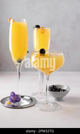 Tre cocktail di mimosa in due flauti e un bicchiere di coupé, ciliegie e arance, una ciotola di ciliegie, stuzzichini e fiori di croco. Foto Stock