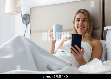 giovane donna sorridente che si posa a letto e usa il telefono mentre beve il caffè del mattino nella camera da letto di casa Foto Stock