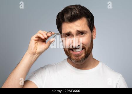 Concetto di depilazione e cura di sé. Bell'uomo di mezza età che bearded tweezing le sopracciglia, levandosi in piedi sopra lo sfondo grigio Foto Stock