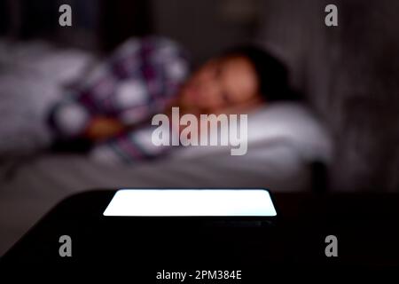 Donna addormentata a letto a casa con il telefono cellulare acceso sul comodino Foto Stock