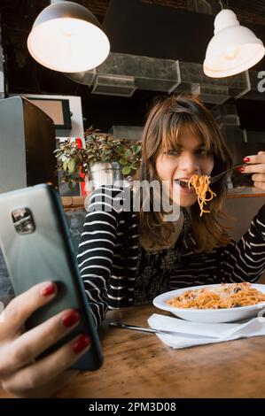 immagine verticale della giovane transgender latina di etnia argentina mangiare spaghetti all'interno di un ristorante e prendere un selfie Foto Stock