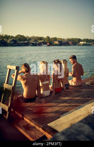 Cinque giovani adulti seduti su un molo di legno in costume da bagno per prendere il sole. Vista posteriore degli amici in vacanza. Vacanza, convivenza, concetto di stile di vita. Foto Stock