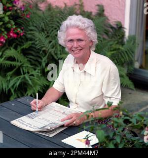 Donna anziana seduta al tavolo da picnic e facendo un puzzle di cruciverba nel giornale Foto Stock