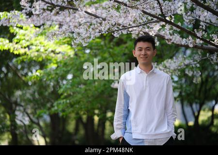 Ritratto frontale del sorriso bel giovane asiatico in camicia casual, mani in tasca. Guardando la macchina fotografica sotto l'albero bianco di sakura Foto Stock