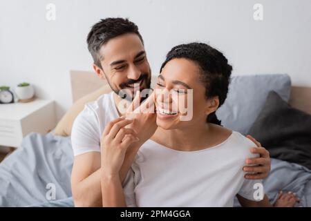 sorridente uomo bearded che tocca faccia della donna afro-americana overjoyed in camera da letto a casa, immagine di riserva Foto Stock