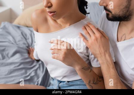 vista ritagliata di uomo bearded che abbraccia una donna afroamericana tatuata seduta in t-shirt bianca in camera da letto, immagine stock Foto Stock