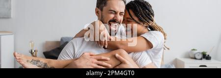 overjoyed coppia interracial in t-shirt bianche abbracciando e ridendo con gli occhi chiusi in camera da letto a casa, banner, immagine stock Foto Stock