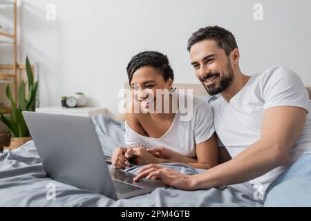 coppia interrazziale gioiosa in t-shirt bianche guardando film sul notebook in camera da letto a casa, immagine stock Foto Stock
