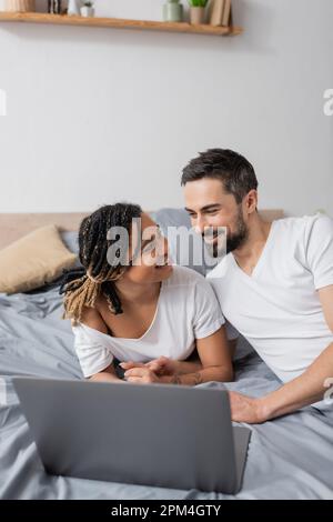 felice coppia multietnica in t-shirt bianche sorridenti l'uno accanto al computer portatile a letto a casa, immagine stock Foto Stock