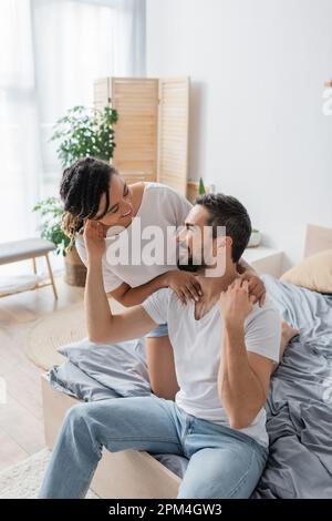allegro coppia interracial in t-shirt bianche che si guardano l'un l'altro mentre hanno divertimento in camera da letto a casa, immagine di scorta Foto Stock