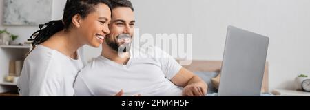 coppia multietnica spensierata in t-shirt bianche guardando film sul portatile a casa, banner, immagine stock Foto Stock