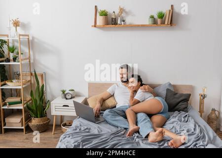 intera lunghezza di spensierata coppia multietnica guardando film sul computer portatile in camera da letto moderna a casa, immagine stock Foto Stock