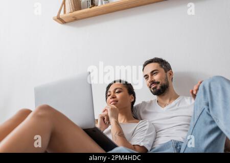 vista dall'angolo basso di una coppia multietnica sorridente che guarda un film sul computer portatile mentre si riposa in camera da letto a casa, immagine di scorta Foto Stock