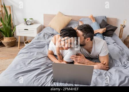 coppia interrazziale overjoyed ridendo con gli occhi chiusi mentre guarda il film sul laptop in camera da letto a casa, immagine di scorta Foto Stock