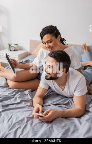 donna afroamericana gioiosa che tiene il tablet digitale vicino sorridente uomo bearded sdraiato con lo smartphone sul letto a casa, immagine di scorta Foto Stock